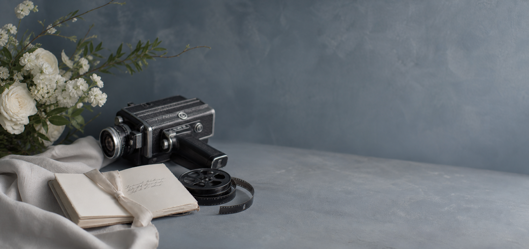 A wedding videography detail scene with a film camera, vow booklet, linen, and white florals on a blue-gray surface.